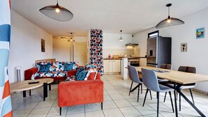 Ein modernes Wohnzimmer mit einer Couch, einem Hocker, einem Esstisch, Stühlen und einer Küchenzeile im Hintergrund.
