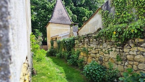 Ein schmaler Steinweg führt zu einem kleinen Gebäude mit Satteldach, umgeben von Grünpflanzen und einer Steinsäule.