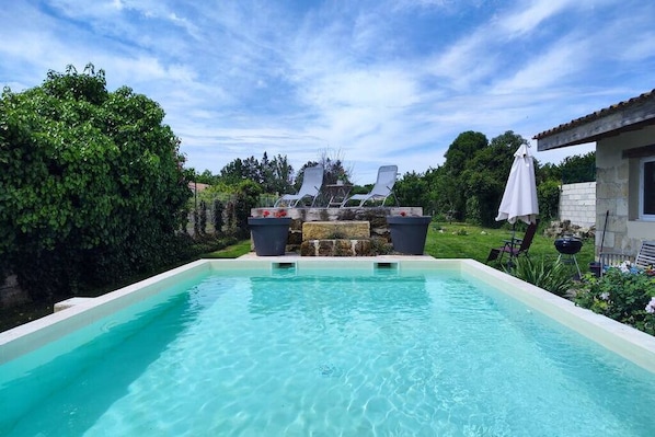 A rectangular swimming pool with clear water, surrounded by greenery and a stone structure in the background.