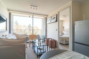 A modern hotel room with a bed, a dining table, a chair, a sofa, a refrigerator, and a large window.