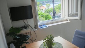 A room with a wooden table, a potted plant, a TV mounted on the wall, and a window with a view of greenery and buildings. - Green House (Mljet)