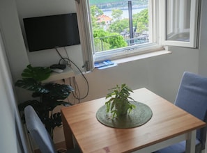 A room with a wooden table, a potted plant, a TV mounted on the wall, and a window with a view of greenery and buildings. - Green House (Mljet)