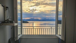 A room with a large window overlooking a body of water and mountains, a chair, and a bed with a towel. - Green House (Mljet)