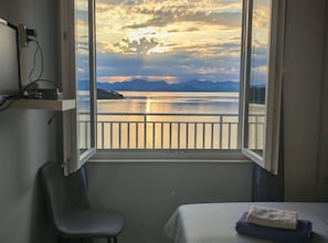 A room with a large window overlooking a body of water and mountains, a chair, and a bed with a towel. - Green House (Mljet)