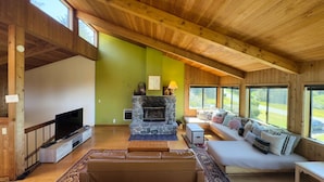 A spacious living room with a stone fireplace, wooden ceiling, large windows, and a cozy seating area.