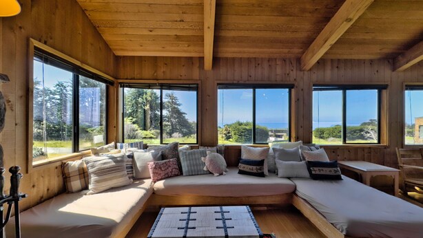 A cozy cabin interior with a large window offering a scenic view, a comfortable seating area with cushions, and a rustic wooden ceiling.