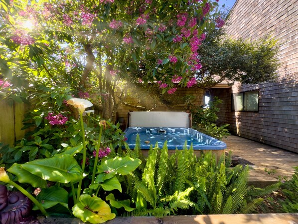 A hot tub surrounded by lush greenery and flowering plants in a backyard setting.