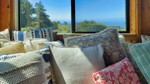 A room with a large window overlooking a forest and ocean, featuring a variety of pillows and a wooden interior.