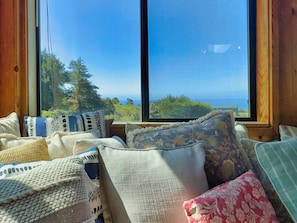 A room with a large window overlooking a forest and ocean, featuring a variety of pillows and a wooden interior.
