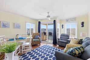 A living room with a ceiling fan, a dining area with a table and chairs, a sofa with cushions, and a view of the beach through large windows. - A Place At The Beach 213 - Coastal Calm Meets Resort Fun! (Atlantic Beach)