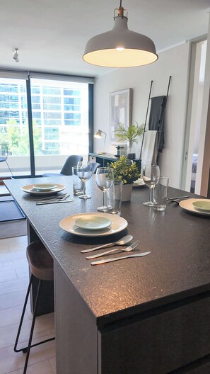 A modern kitchen with a dark countertop, a hanging pendant light, and a view of the city through large windows. - HOM TOP1 | Las Condes | Parque Arauco | Parking (Las Condes)