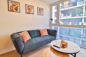 Ein modernes Wohnzimmer mit einem grauen Sofa, gemusterten Kissen, einem runden Couchtisch, einer Fernbedienung und einem Fenster mit Blick auf ein Gebäude.