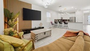 Uma sala de estar moderna com televisão de tela plana, uma parede de destaque em madeira, um espaço para refeições com banquetas de bar e uma cozinha ao fundo.