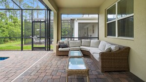 Um alpendre coberto com um sofá de vime, uma mesa de centro de vidro e vista para uma piscina e vegetação.
