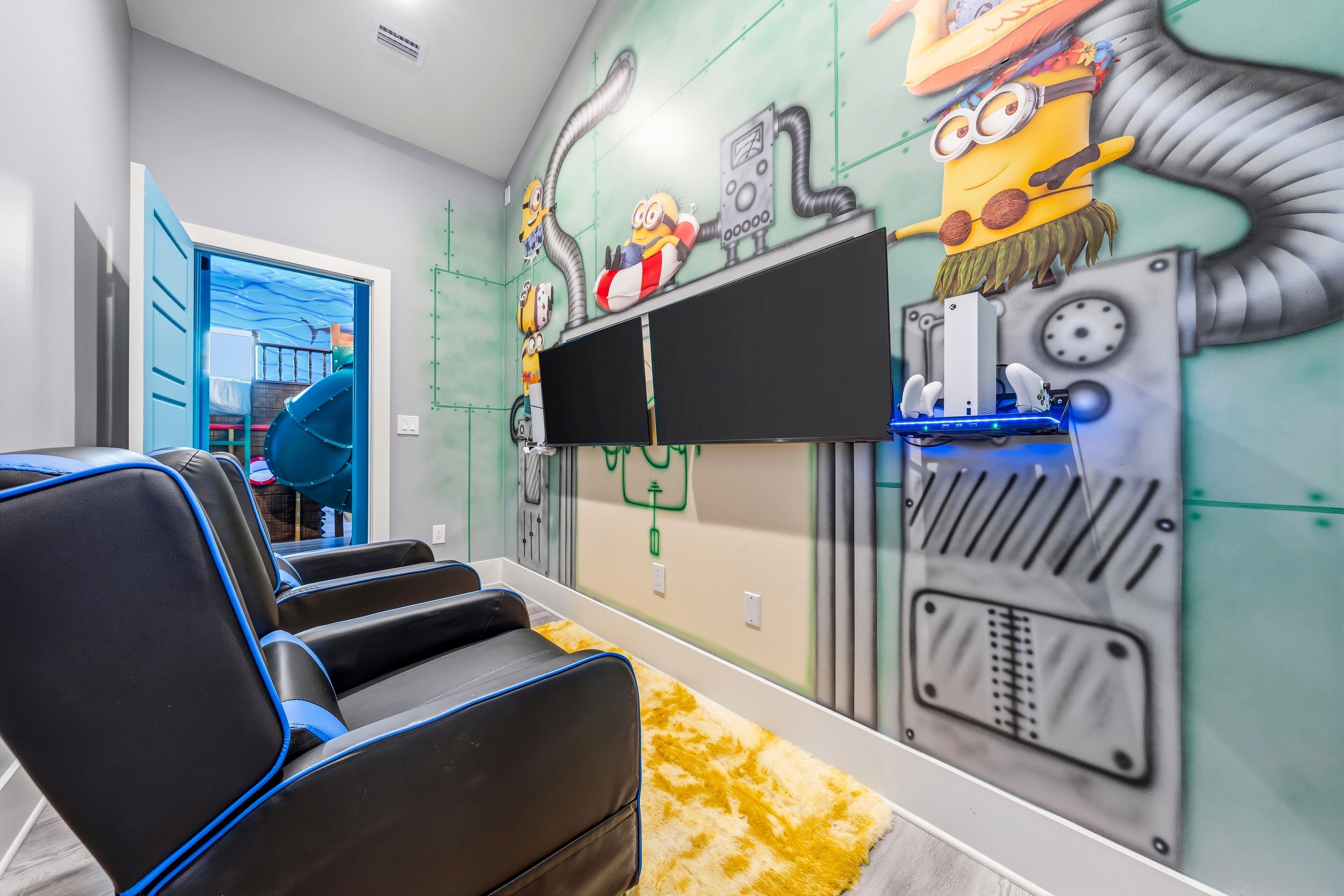 Une pièce avec un téléviseur fixé au mur, un coin salon avec des fauteuils noirs et bleus, et une fresque murale représentant des personnages de dessins animés.