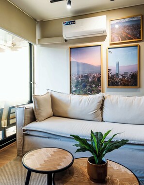 Un salon moderne avec un canapé, une table ronde en bois, une plante en pot et deux tableaux représentant des paysages urbains accrochés au mur.