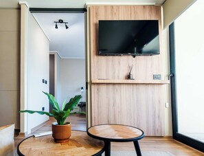 Un salon moderne avec un téléviseur à écran plat fixé sur un mur en bois, une table ronde en bois, une plante en pot et un couloir menant à une autre pièce.