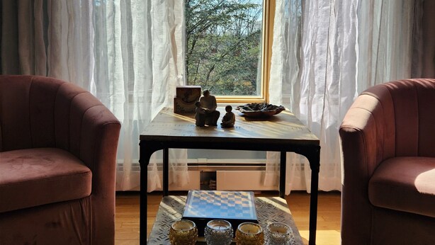 A room with two armchairs, a small table with chess pieces and a bowl, and a view of trees through sheer curtains.