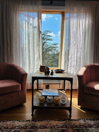 A room with two armchairs, a small table with chess pieces and a bowl, and a view of trees through sheer curtains.. Welcome to The Manse on the Hill 2-Story Retreat 3 Beds Home Parking PetFriendly