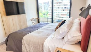 Ein Hotelzimmer mit Bett, Wandfernseher, Sessel und Blick auf Gebäude durch das Fenster.
