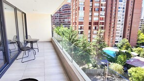 A balcony with a table and chairs, a glass railing, and a view of a high-rise building and a swimming pool. - HOM TOP1 | 97m2 | Parking | Parque Arauco | 1D1B (Las Condes)