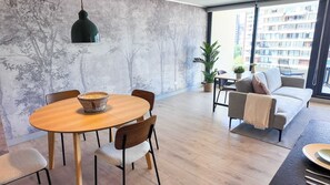 Ein modernes Wohnzimmer mit einem runden Holztisch, zwei Stühlen, einem Sofa, einer hängenden Pendelleuchte und einem großen Fenster mit Blick auf Gebäude.