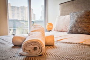 Una cama bien hecha con toallas enrolladas, una almohada y una lámpara de noche en una habitación de hotel con vistas al exterior.