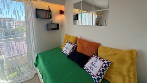 Ein Schlafzimmer mit grüner Bettdecke, mehreren Kissen, einer kleinen Ablage mit Büchern und einem Dekorationsgegenstand, einem wandmontierten Spiegel und einem Fenster mit transparenten Vorhängen.
