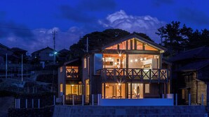 Una casa de madera de dos pisos con balcón, ventanas amplias y un interior visible con muebles.
