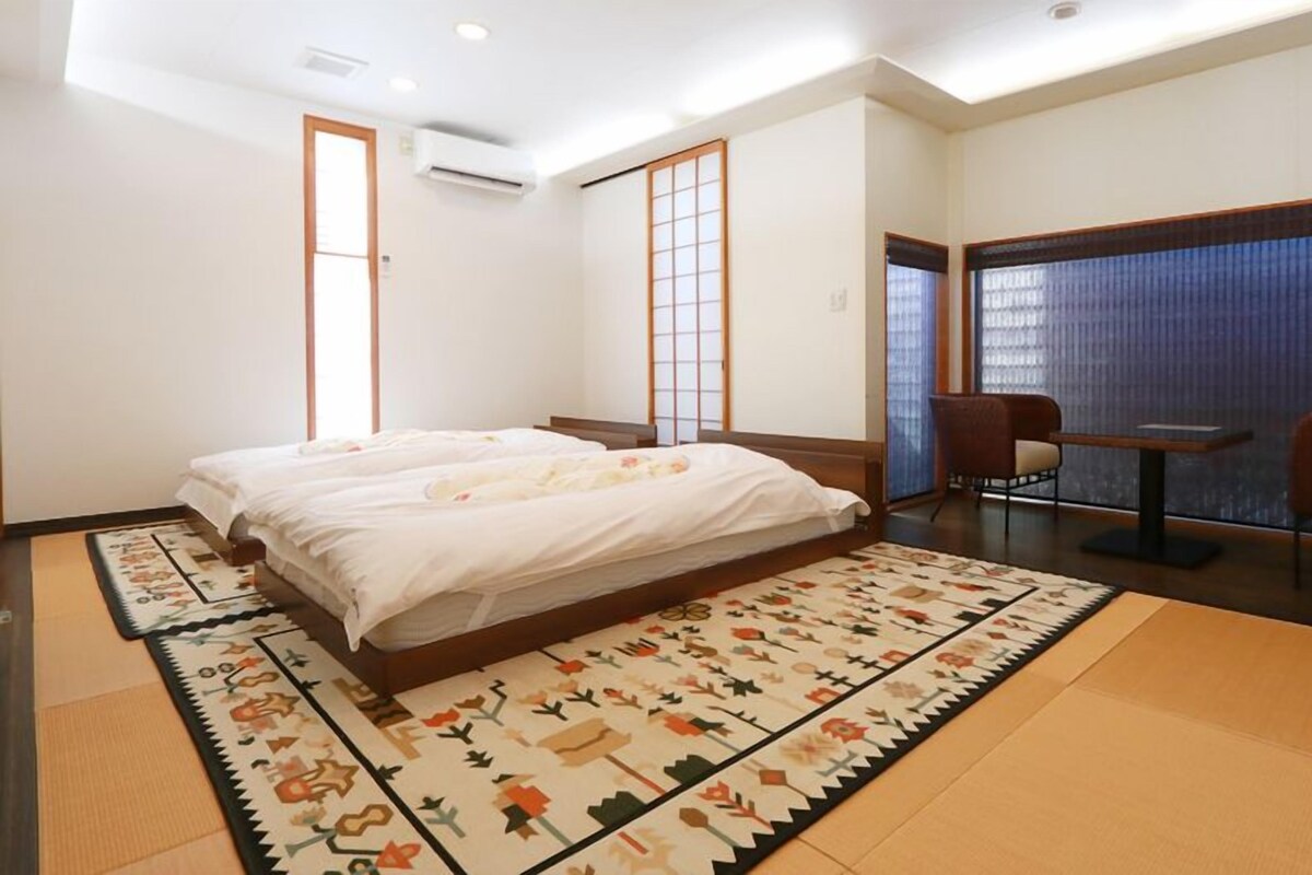 Une chambre d’hôtel avec un lit, une petite table, deux chaises, une fenêtre avec des stores et un paravent japonais traditionnel en papier.