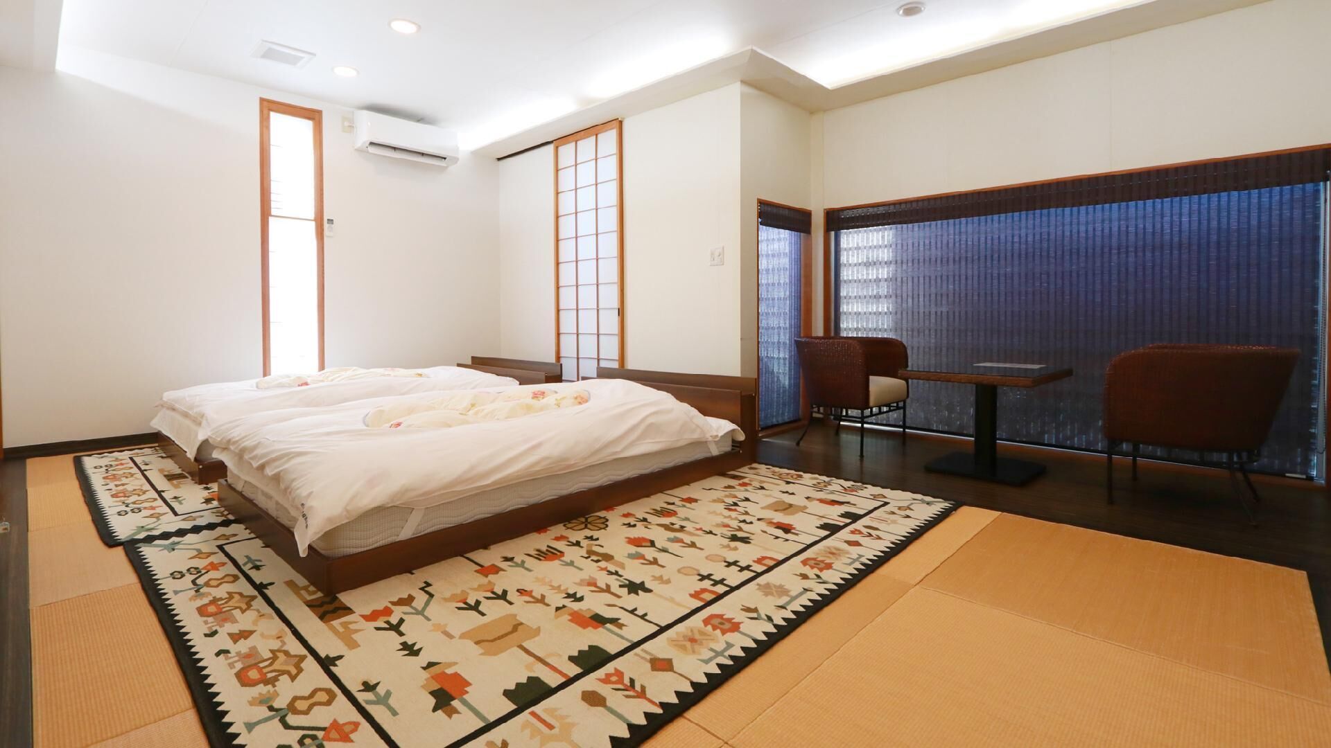Ein Hotelzimmer mit einem großen Bett, zwei Stühlen, einem kleinen Tisch, einer Tatami-Matte und einer Schiebetür.