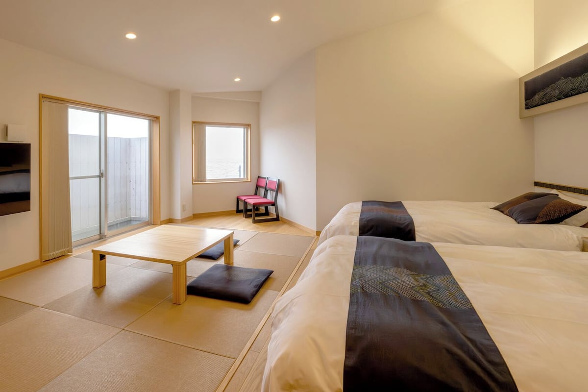 A hotel room with two beds, a small table, a chair, and sliding doors leading to a balcony.