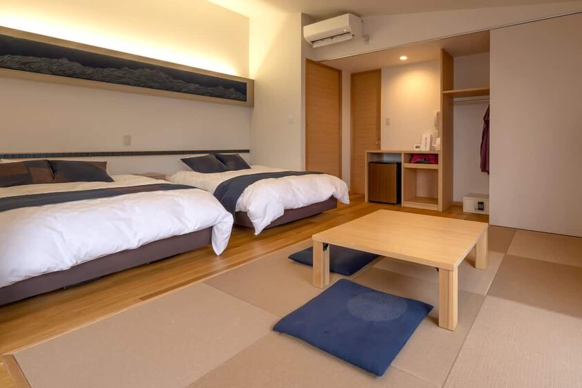 A hotel room with two beds, a small table, a wardrobe, and an air conditioning unit.