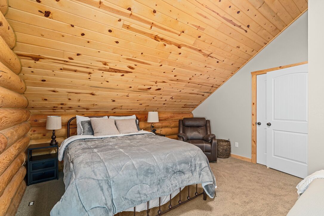 A log cabin bedroom with a sloped ceiling, a bed with a grey quilt, a leather chair, and a white door.