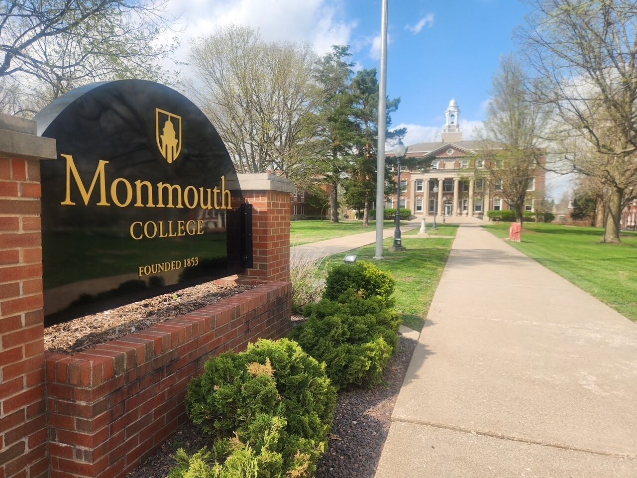 A brick entrance with a sign for Monmouth College, founded in 1853, and a pathway leading to a building with columns.