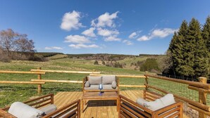 Une terrasse en bois avec un canapé et deux fauteuils, donnant sur un paysage verdoyant parsemé d’arbres et sous un ciel dégagé.