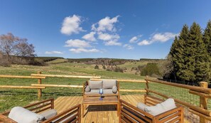 Eine Holzterrasse mit einer Couch und zwei Stühlen, mit Blick auf eine grüne Landschaft mit Bäumen und blauem Himmel.