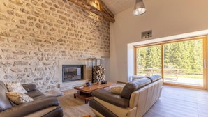 Un salon aux murs en pierre, avec une cheminée, des canapés en cuir et une grande fenêtre donnant sur la forêt.