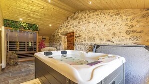 Un espace spa doté d’un jacuzzi, de murs en pierre, d’un plafond en bois et d’une véranda en verre où sont disposées des plantes.