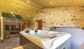 Wellnessbereich mit Whirlpool, Steinwänden, Holzbalkendecke und einem Glasbereich mit Pflanzen.