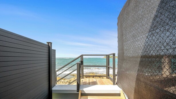 Una escalera de madera que baja a una playa, con baranda de metal, una pared gris y una malla protectora.