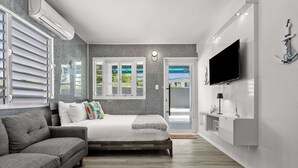 Un dormitorio moderno con cama, sofá gris, televisor y vistas al verdor por la ventana.