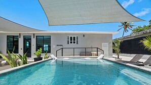 Una piscina con sillas de descanso y una zona sombreada, rodeada por un edificio con grandes ventanales y una puerta.