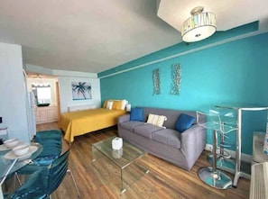 A room with a bed, a sofa, a glass coffee table, and a dining area with a table and chairs.