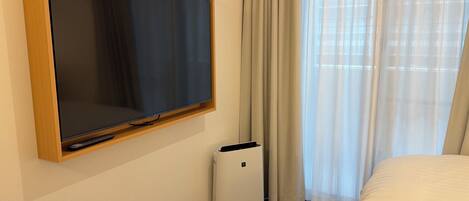 A hotel room with a mounted TV, a standing heater, a bed, and a window with curtains.