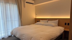 A hotel room with a bed, an air conditioning unit, a bedside table, and a window with curtains. - reside global kumamoto (Kumamoto)