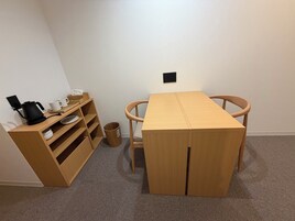 A small, neatly arranged room with a wooden table, chairs, a shelf with cups and a kettle, and a trash bin.