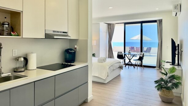 A modern hotel room with a kitchenette, a bed, a balcony with a view of the sea, and a dining area.