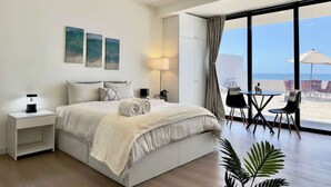 A modern hotel room with a large bed, bedside table, lamp, and a view of the beach through large windows.