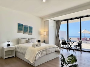 A modern hotel room with a large bed, bedside table, lamp, and a view of the beach through large windows. - Little Pearl Sea View Studio (San Antonio del Mar)